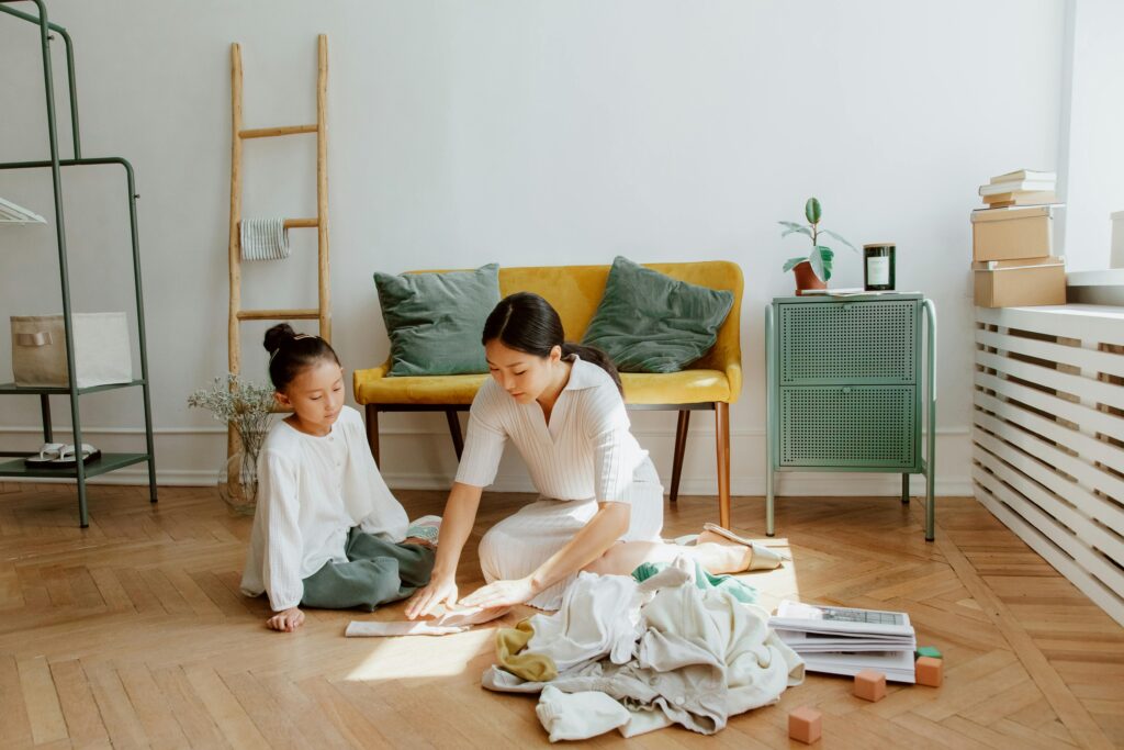 A mother and daughter folding clothes together on the floor in a cozy, sunlit living room.