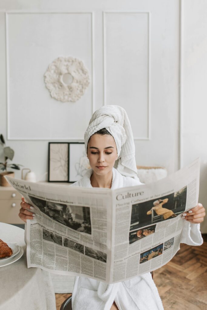 Woman in a bathrobe with head towel reading a newspaper, enjoying a quiet morning indoors.