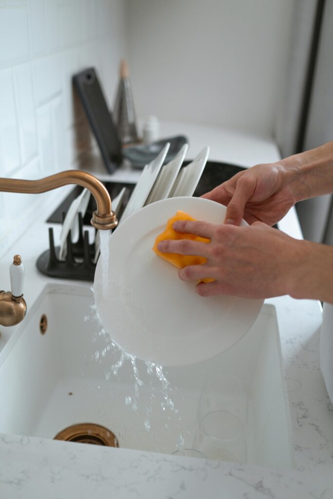 Hands washing a white dish in a modern kitchen sink with a yellow sponge.