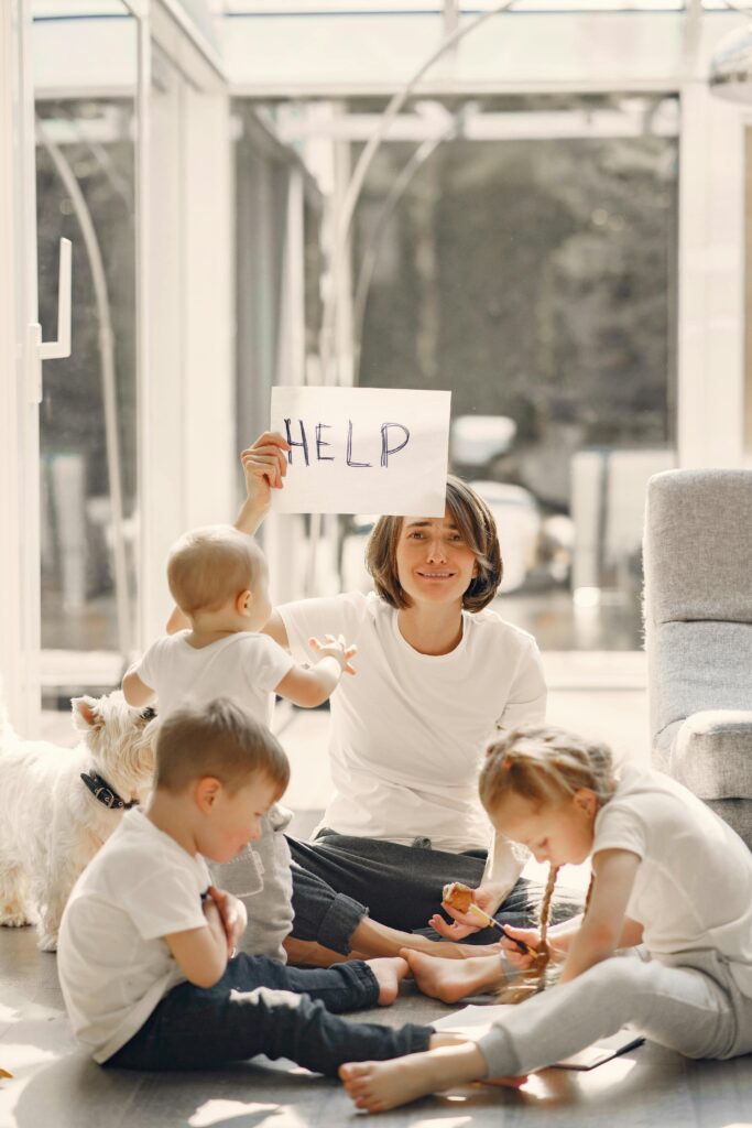 A mother holds a 'HELP' sign while sitting with her children and a dog in a sunlit living room.